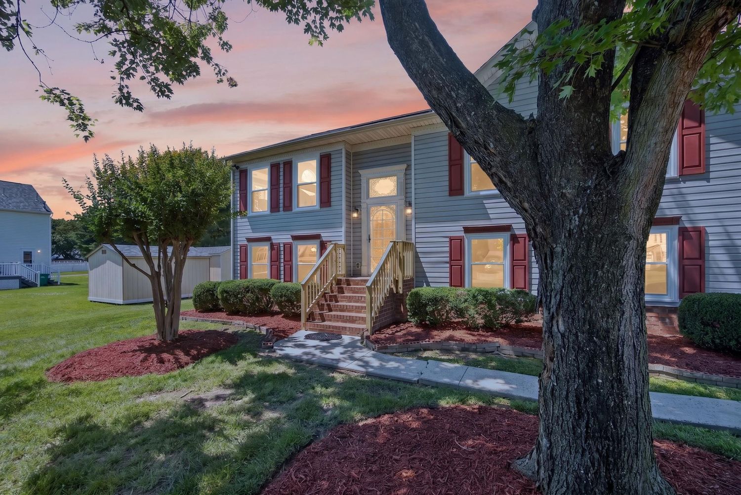 Two-story gray townhome with red shutters, lush landscaping, and a welcoming entrance at sunset.