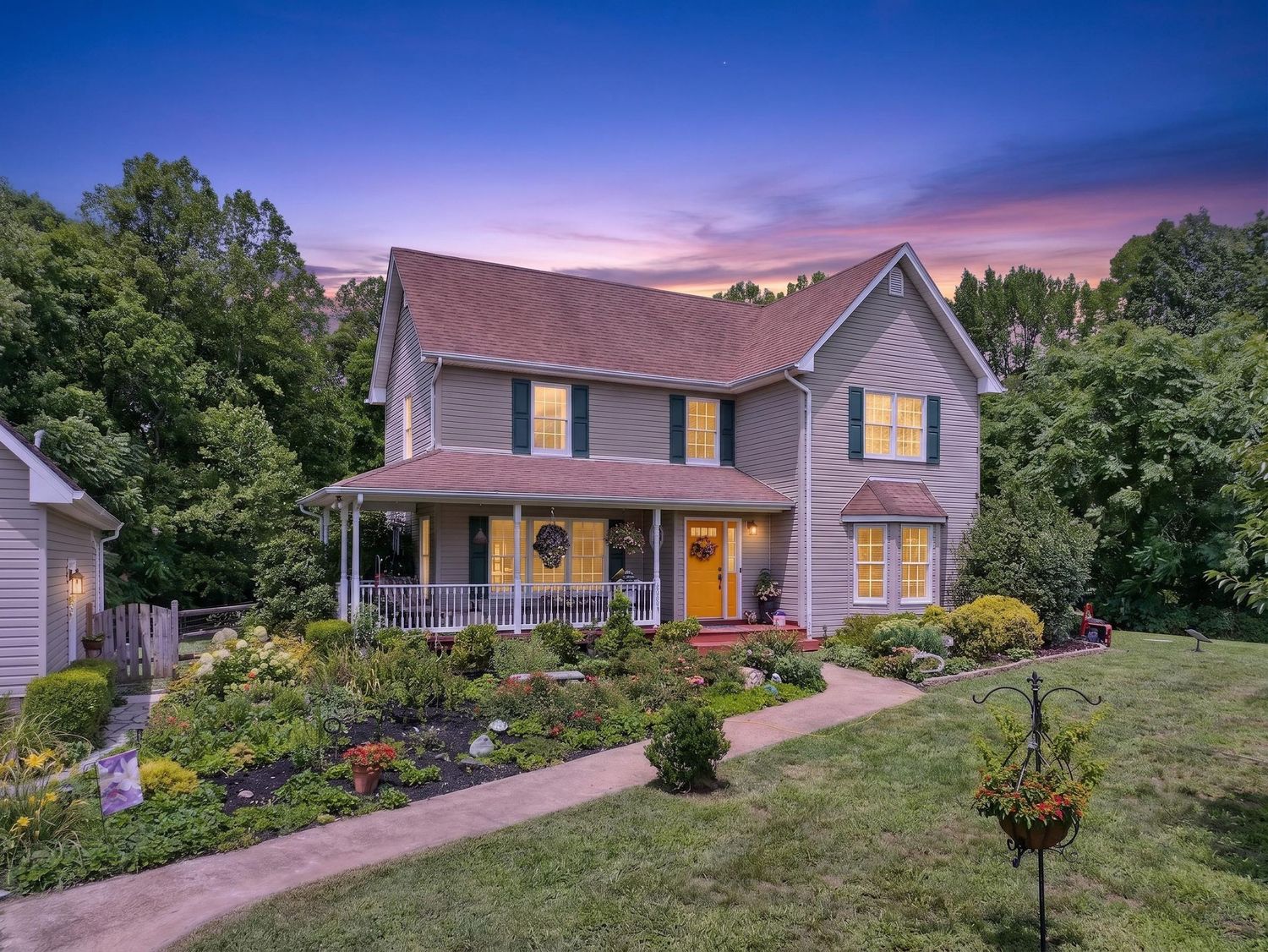 Two-story colonial home with wrap-around porch, colorful garden, and warm glowing windows at twilight.