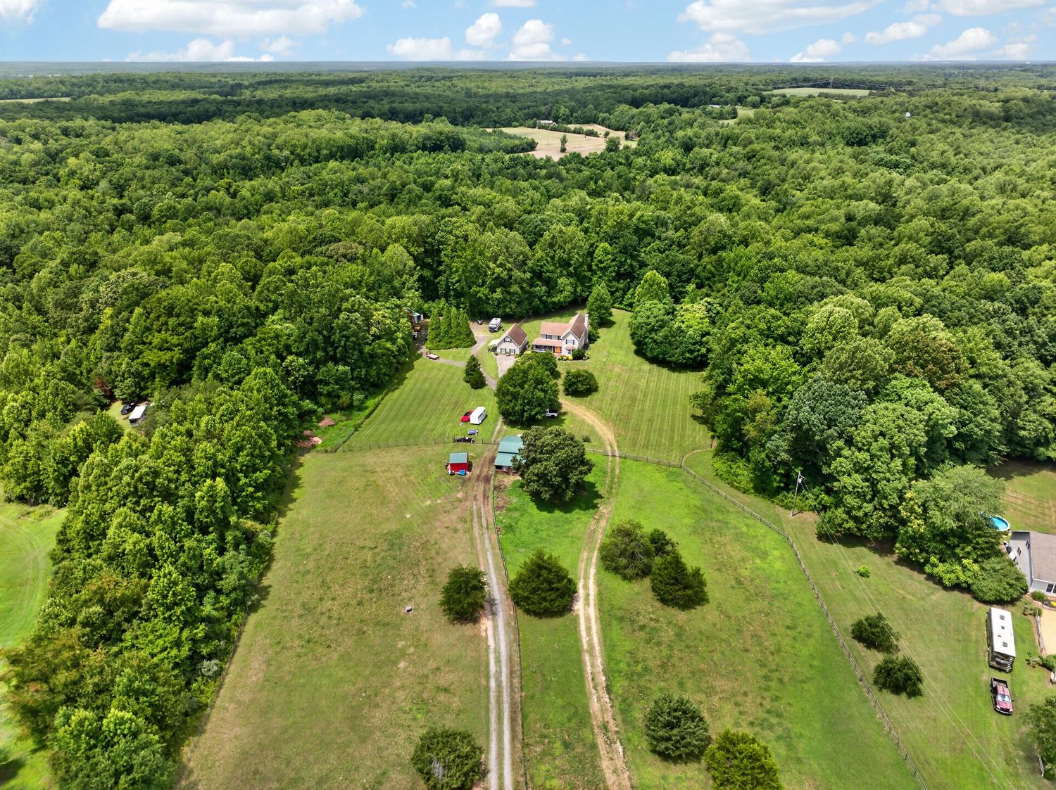 Aerial view of a rural property with a home surrounded by lush green trees and open fields with a long driveway.