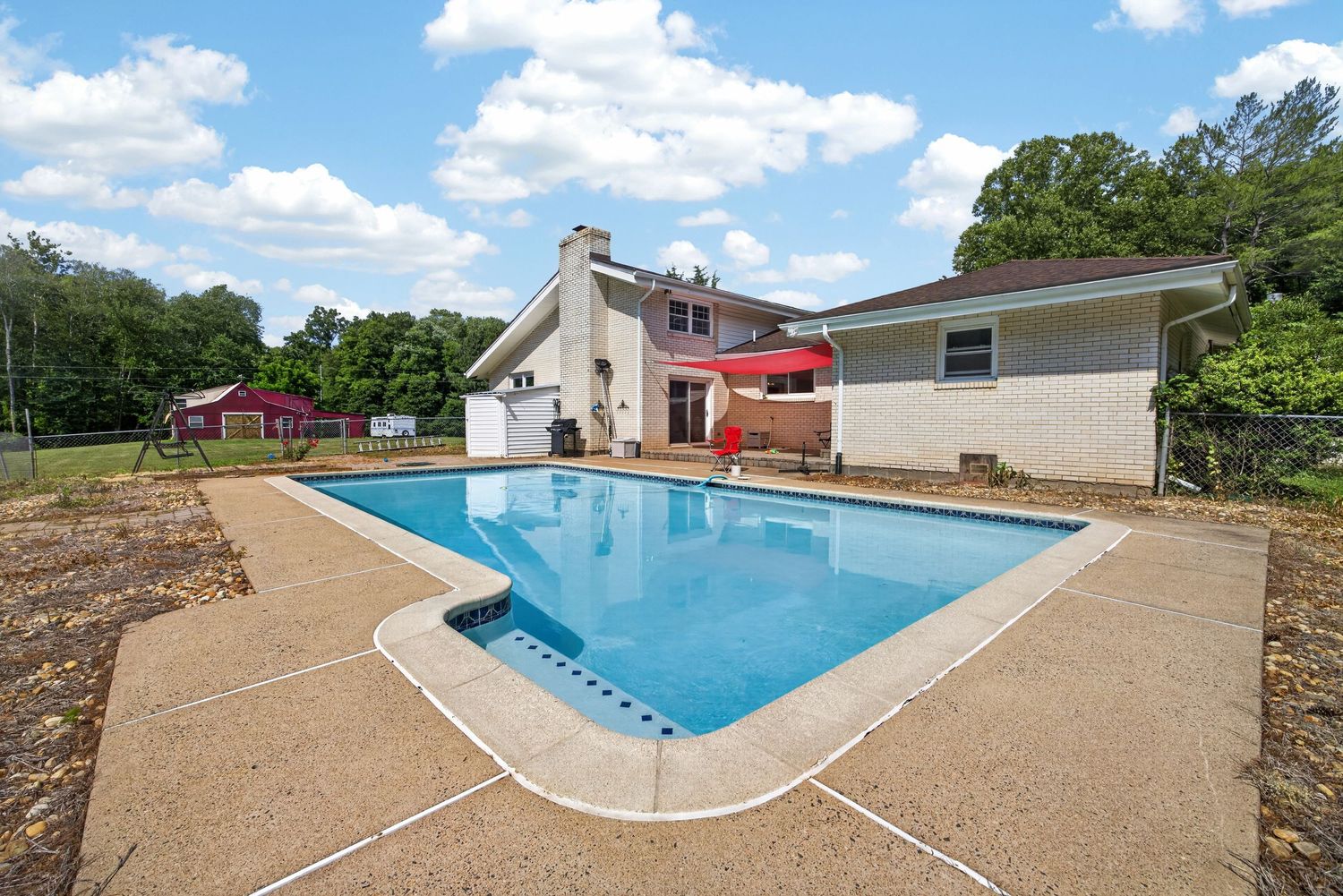 Outdoor rectangular swimming pool with concrete deck beside a two-story house, surrounded by lush green trees.