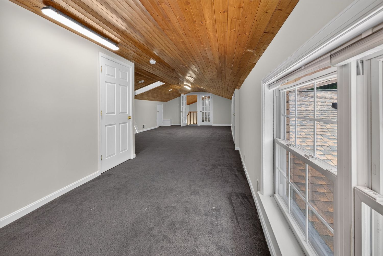 Spacious attic room with vaulted wood ceiling, gray carpet, white walls, and natural light from large windows.