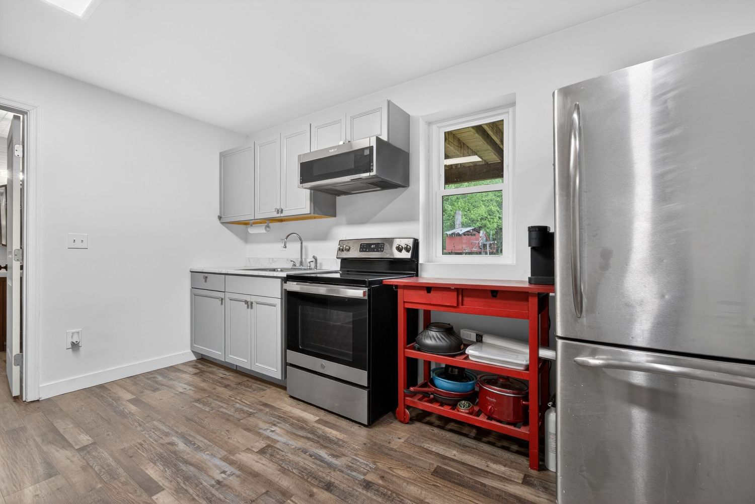 Modern kitchen with stainless steel appliances, gray cabinets, hardwood floors, and a red utility cart near the window.