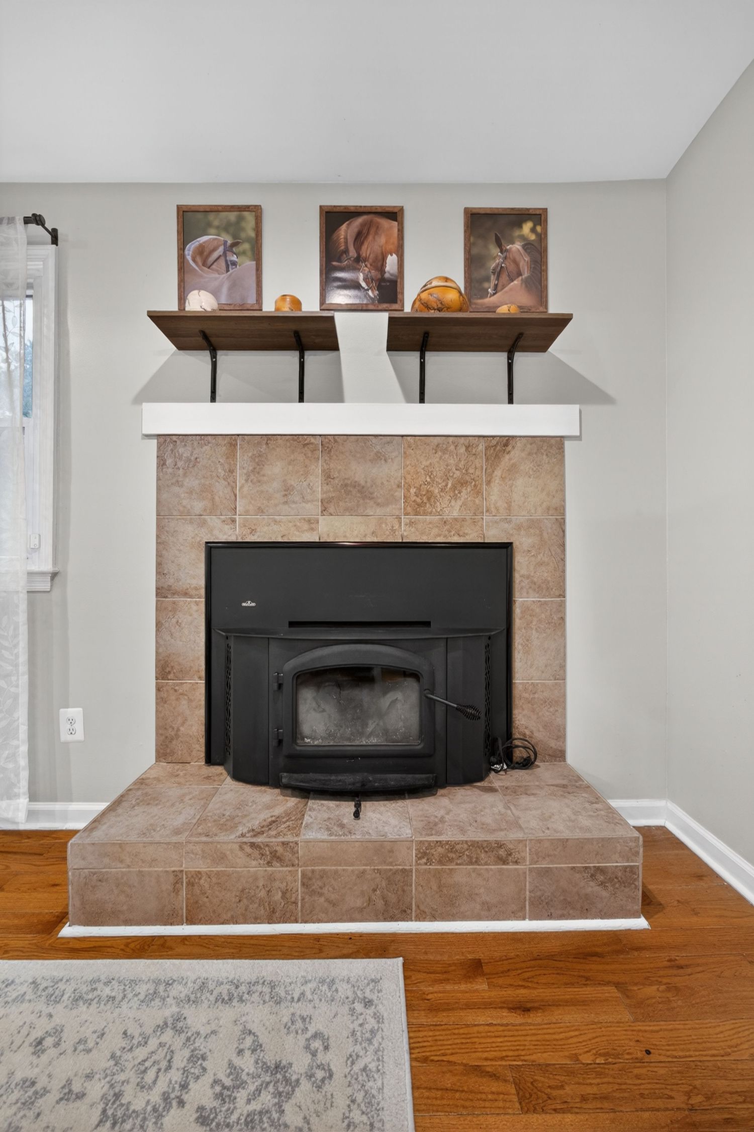 Tiled fireplace with black wood-burning insert, white mantel shelf with framed photos, and hardwood floors in a cozy living room.