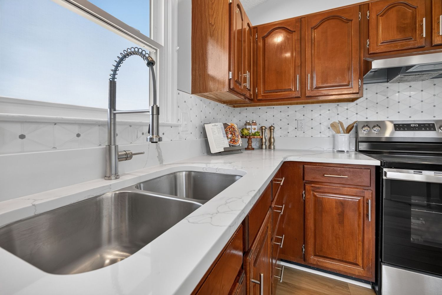 Modern kitchen with cherry wood cabinets, stainless steel double sink, quartz countertops, and electric range with white tile backsplash.