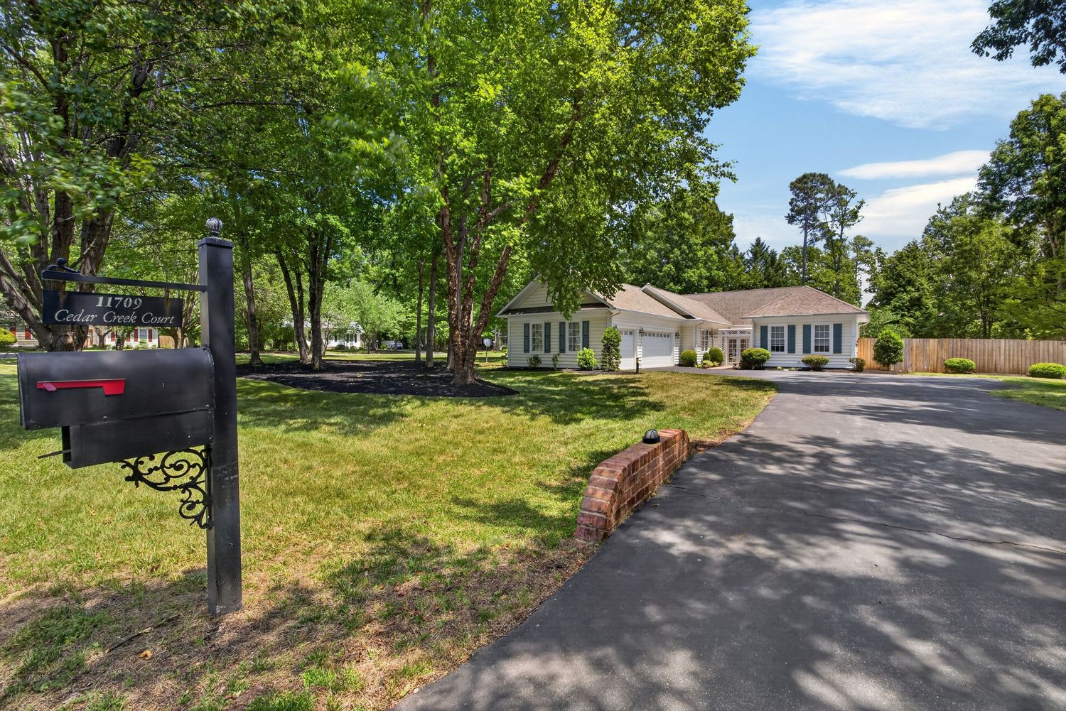Tree-lined driveway leads to a white ranch-style home with lush green lawn and black mailbox in foreground.