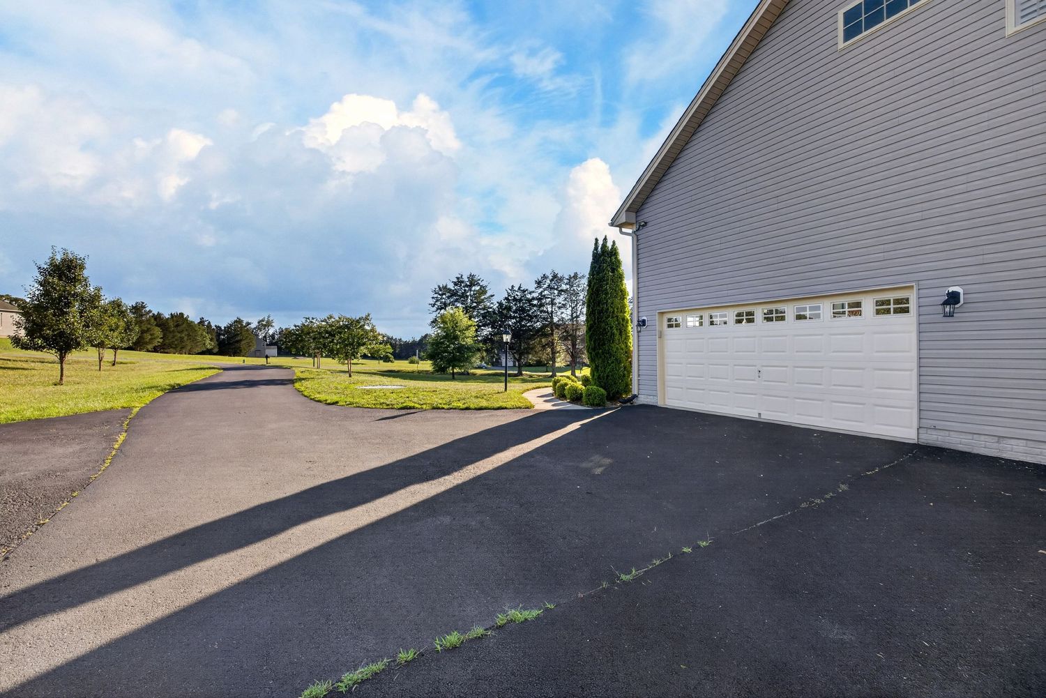 Two-car garage with white doors on a home with a paved driveway surrounded by lush green lawn and trees under a blue sky.