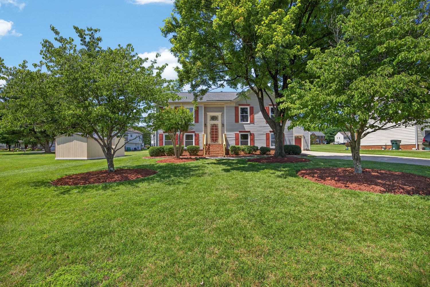 Brick colonial home with lush green lawn, mature trees, and manicured red mulch landscaping on a sunny day.