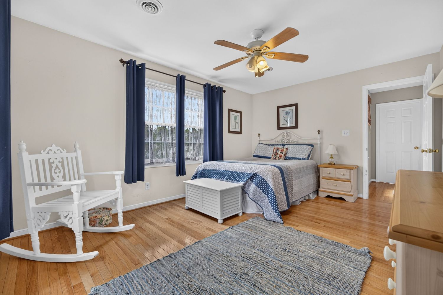 Bright bedroom with hardwood floors, blue curtains, ceiling fan, rocking chair, and a neatly made bed with blue accents.