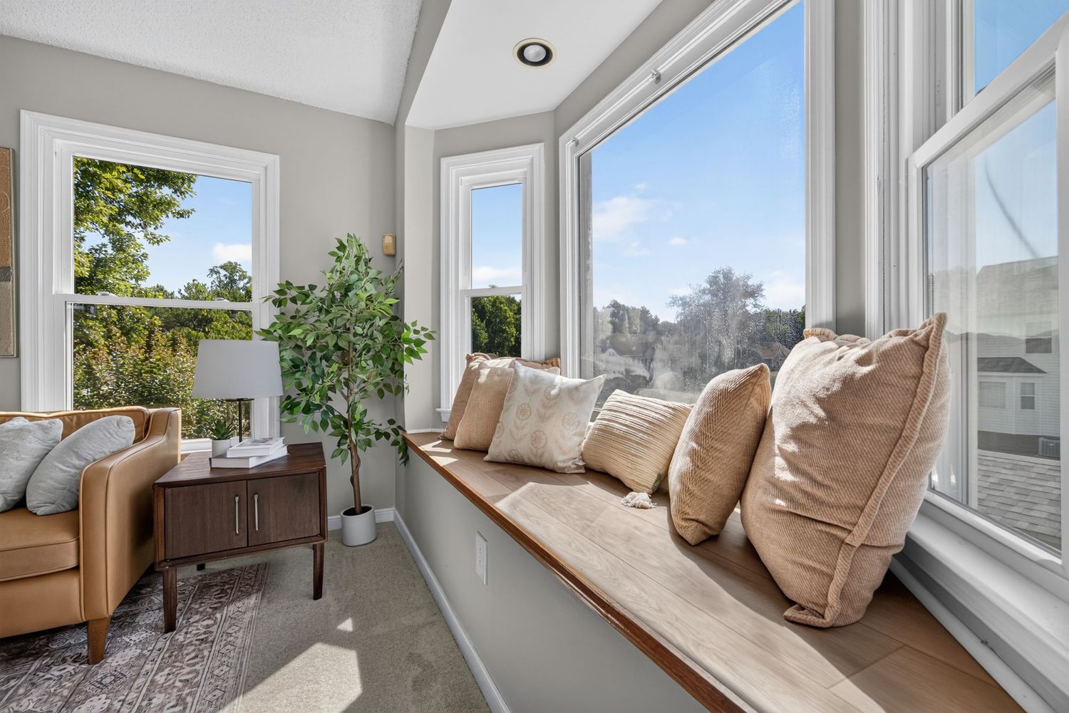 Bright sunroom with built-in window seat, plush pillows, large windows, and a leather armchair beside a potted plant.