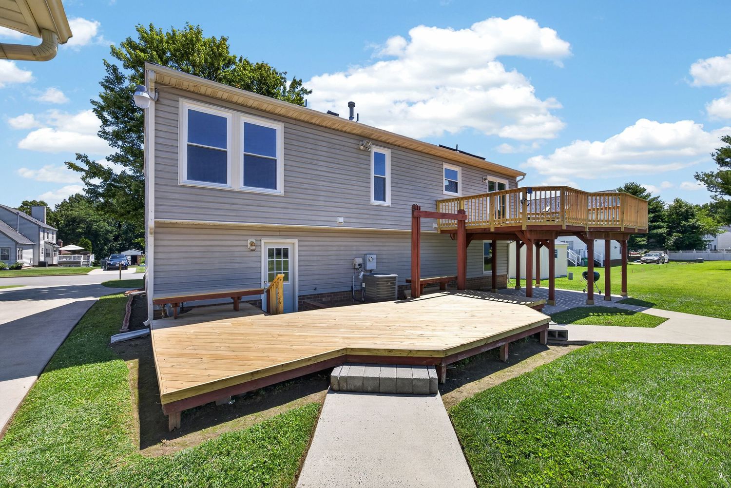 Suburban home with large wooden deck, upper balcony, green lawn, and blue sky in a residential neighborhood.