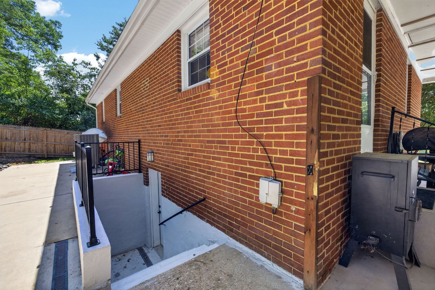 Brick exterior wall of a residential building with a basement entrance, metal railing, and utility equipment on a concrete path.