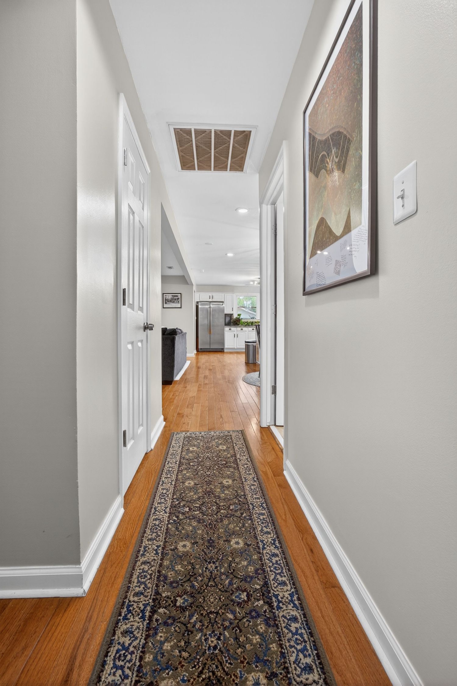 Elegant hallway with hardwood floors, a decorative runner rug, white trim, and framed wall art leading to a modern kitchen.