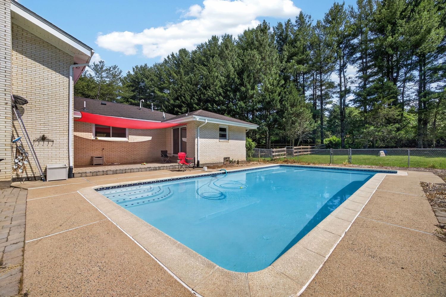 Backyard rectangular swimming pool with concrete patio, red umbrella, pool house, and tall trees in background.