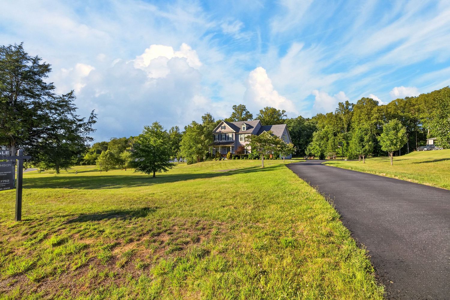 Winding asphalt driveway leading to a large country home surrounded by lush green lawns and mature trees under a blue sky.