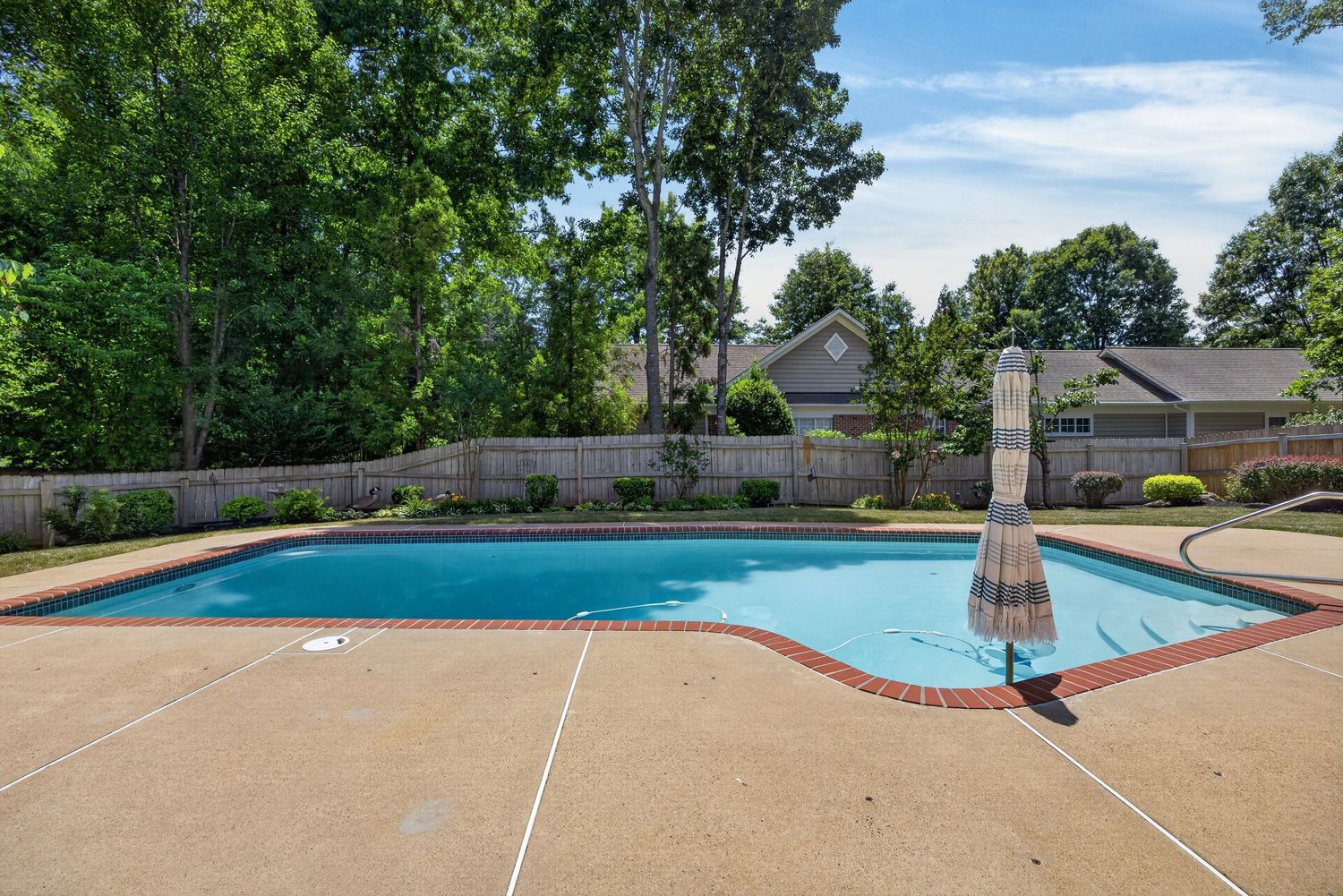 Backyard swimming pool with brick border, concrete patio, and lush green trees surrounding a fenced yard.