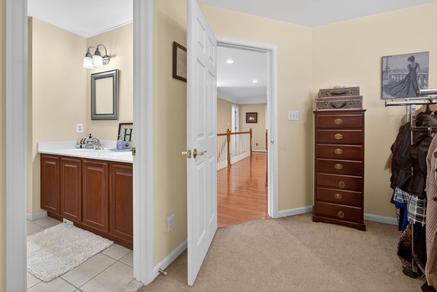 Bright hallway leading to a bathroom with cherry wood vanity and a bedroom with tall dresser and carpeted floors.