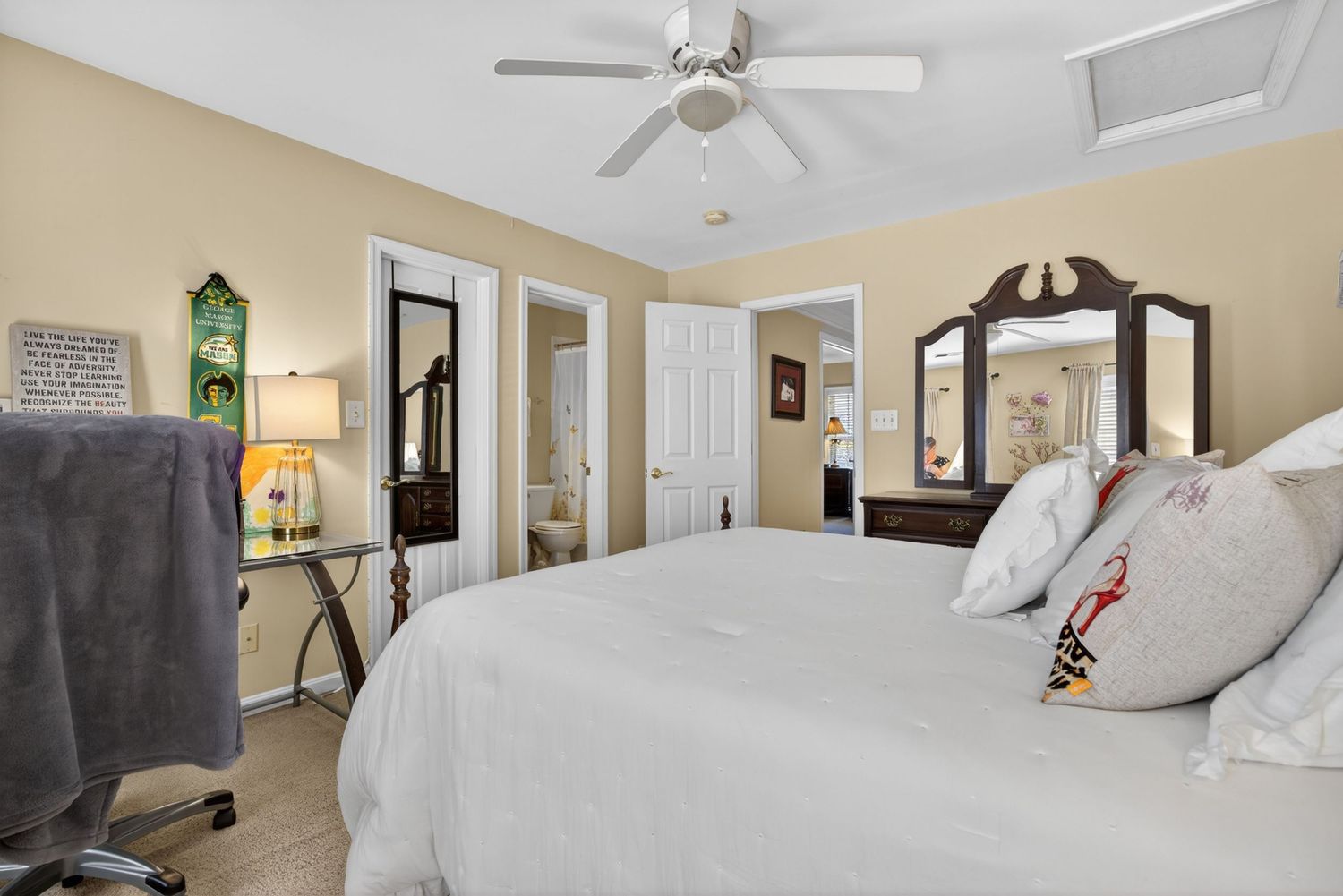 Bright bedroom with white bedding, ceiling fan, mirrored dresser, and doors leading to closet and ensuite bathroom.