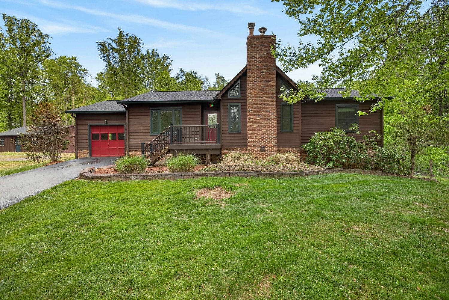 Brown ranch-style home with red garage door, brick chimney, and lush green lawn surrounded by mature trees.