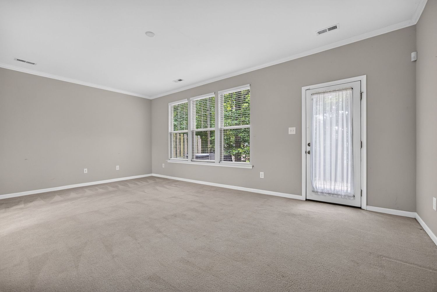 Bright empty living room with gray carpet, crown molding, large windows, and a glass door leading outside.