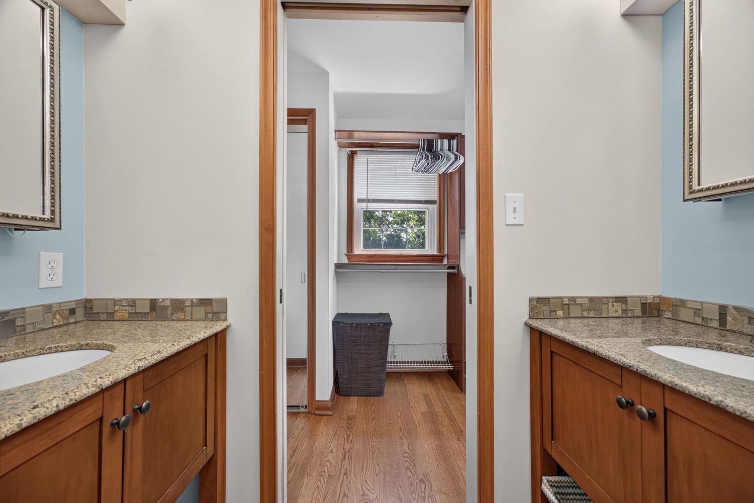 Double vanity bathroom with granite countertops, wood cabinets, and a hallway leading to a bright window beyond.