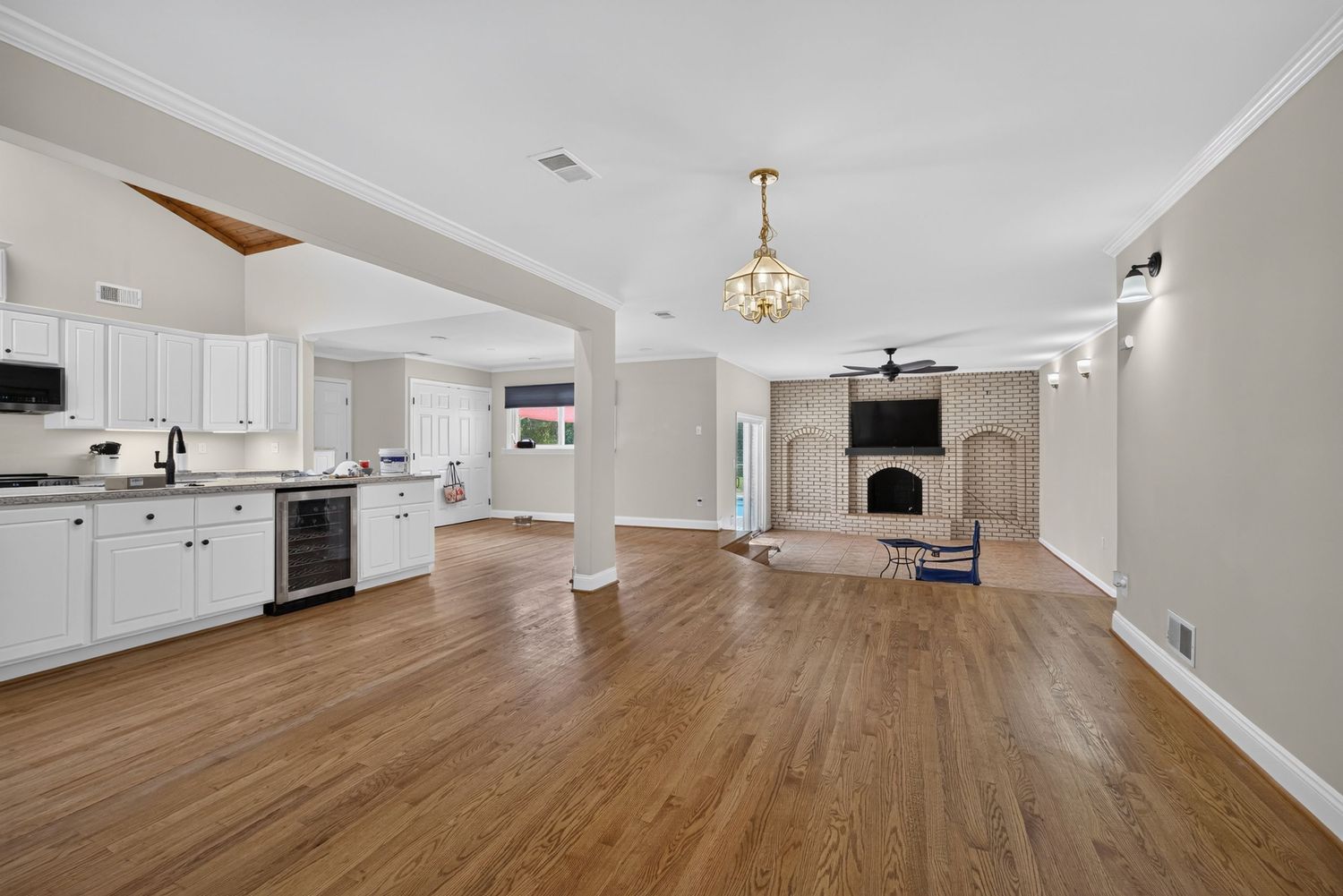 Open-concept living area with hardwood floors, white kitchen, brick fireplace, and elegant chandelier in a bright home.
