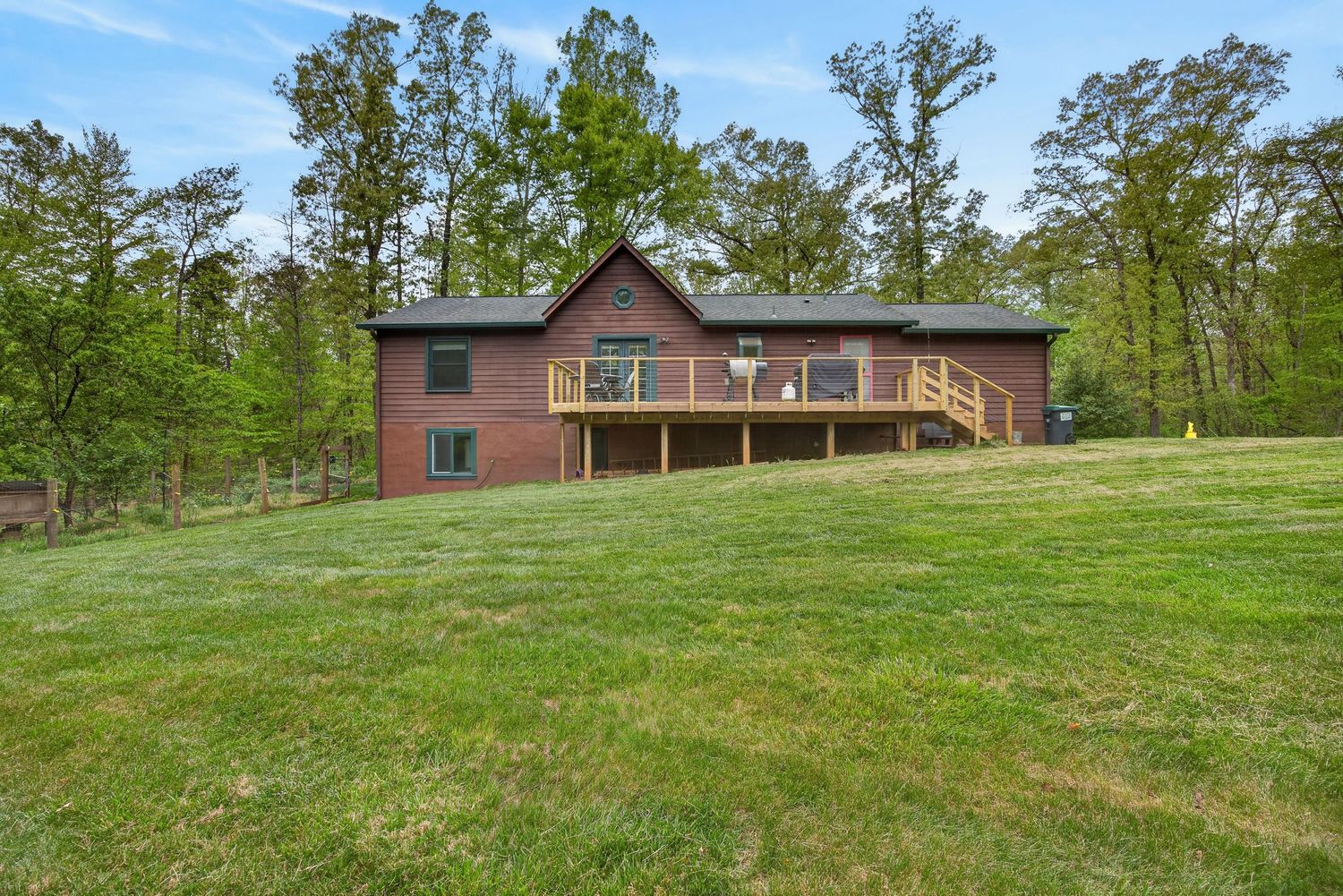 A rustic two-story cabin with a wooden deck sits on a lush green hillside surrounded by tall trees.