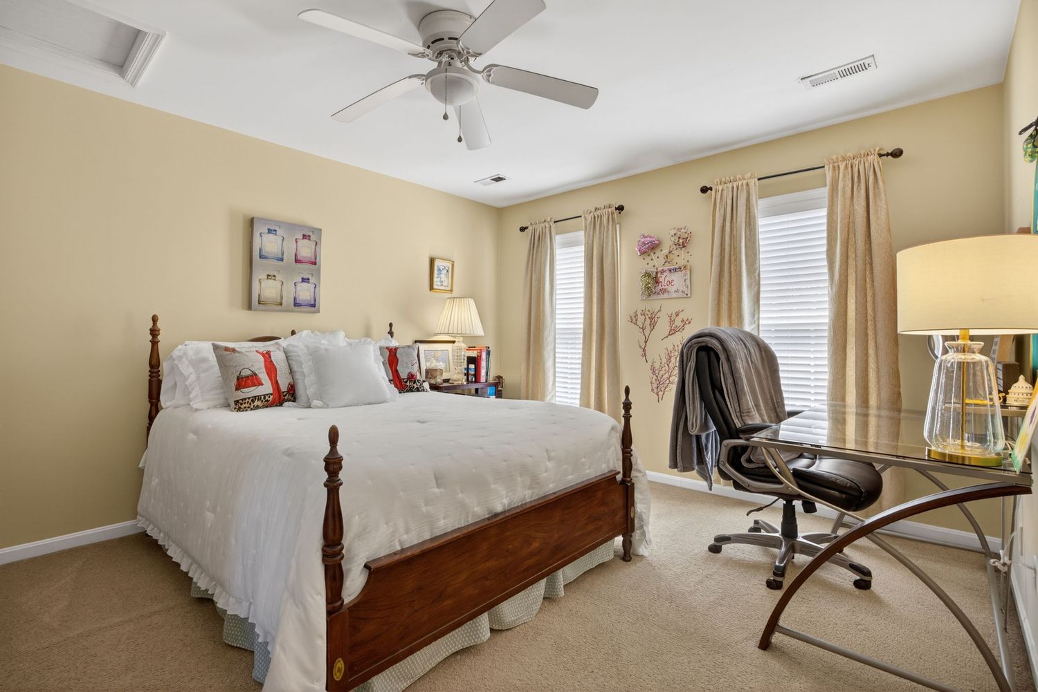 Bright bedroom with wood four-poster bed, ceiling fan, beige carpet, and a desk area near curtained windows.