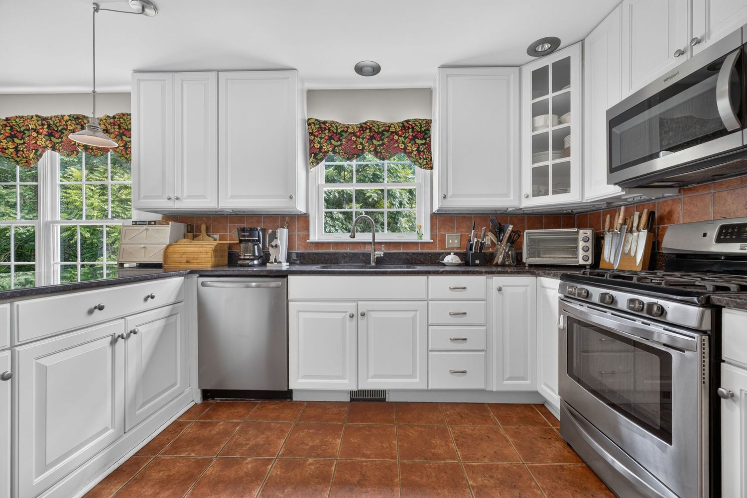 White kitchen with stainless steel appliances, glass-front cabinets, dark countertops, and terracotta tile flooring near a sunny window.