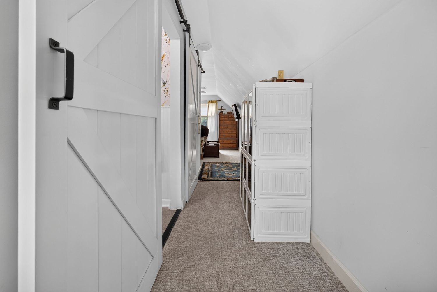 Narrow hallway with white barn door, built-in shelving, and carpet leading to a bright room beyond.