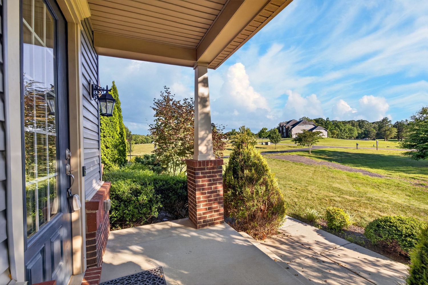 Covered front porch with brick column overlooking a lush green lawn and landscaped shrubs under a partly cloudy sky.