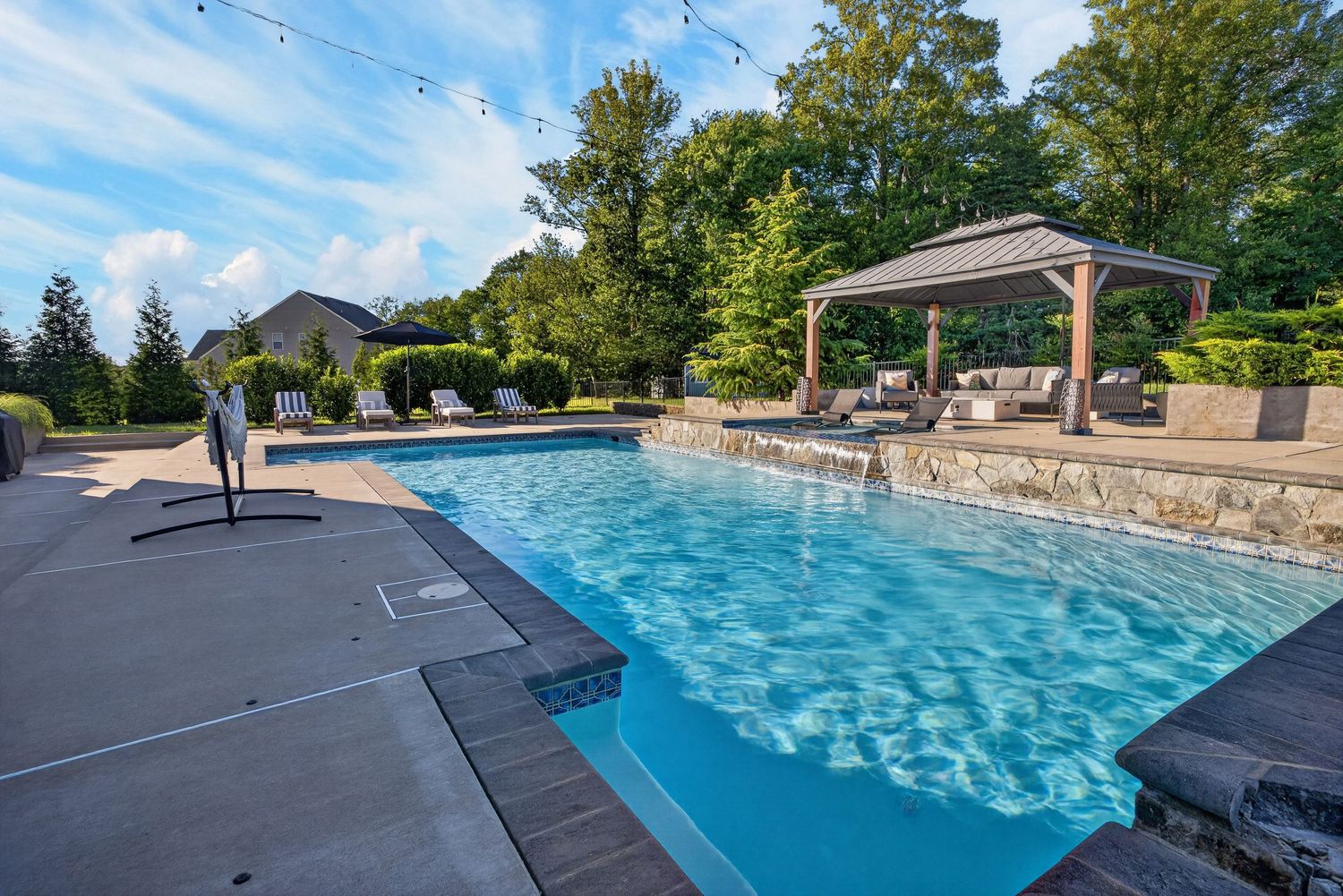 Luxurious backyard pool with crystal-clear water, pergola seating area, and lush green landscaping on a sunny day.