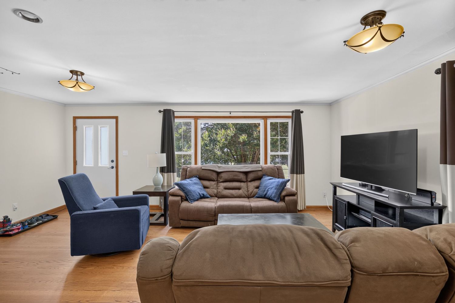Cozy living room with brown leather sofas, blue armchair, hardwood floors, large window, and flat-screen TV on wooden stand.