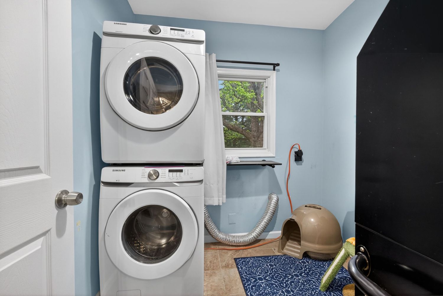 Stacked white washer and dryer unit in a laundry room with blue walls, tile floor, and a window.