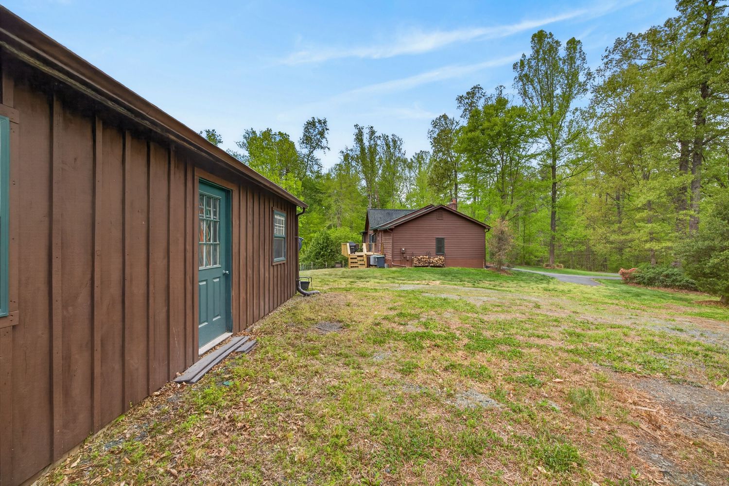 Brown wooden cabin with green door beside a grassy yard and a second cabin visible in the background surrounded by trees.