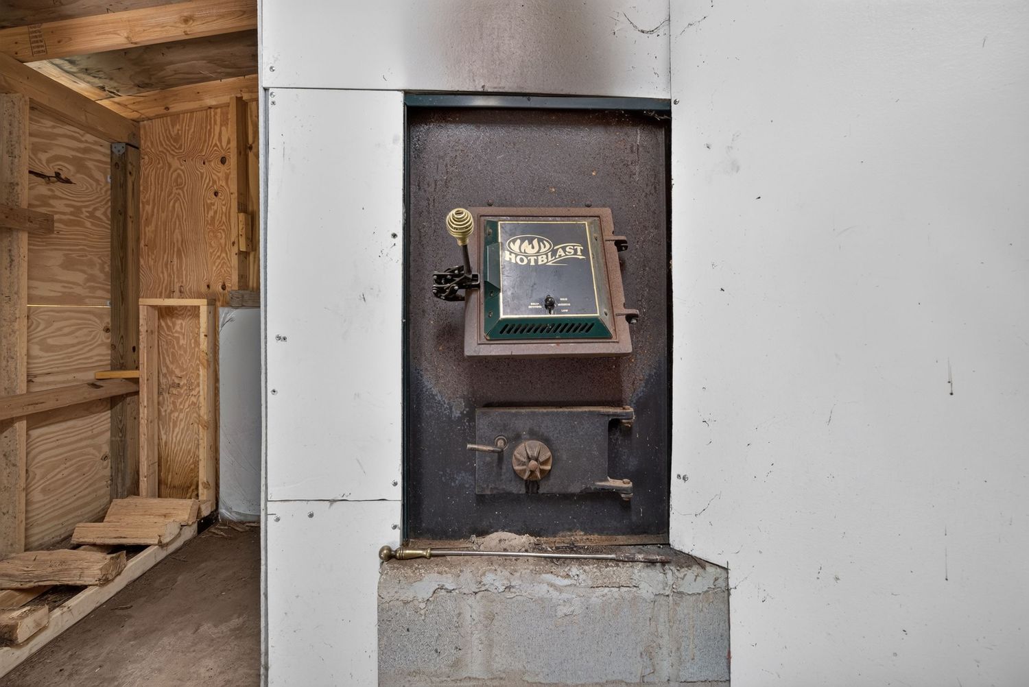 Vintage cast iron furnace door built into a concrete base in a basement utility room with wood framing visible.