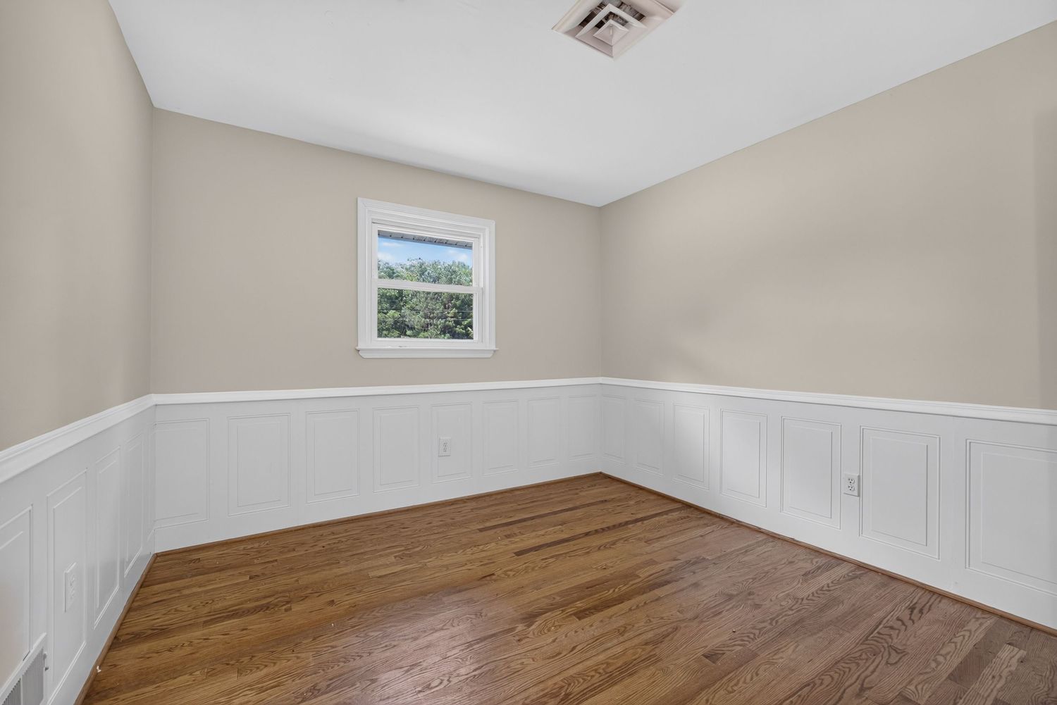 Empty room with hardwood floors, white wainscoting, beige walls, and a single window letting in natural light.