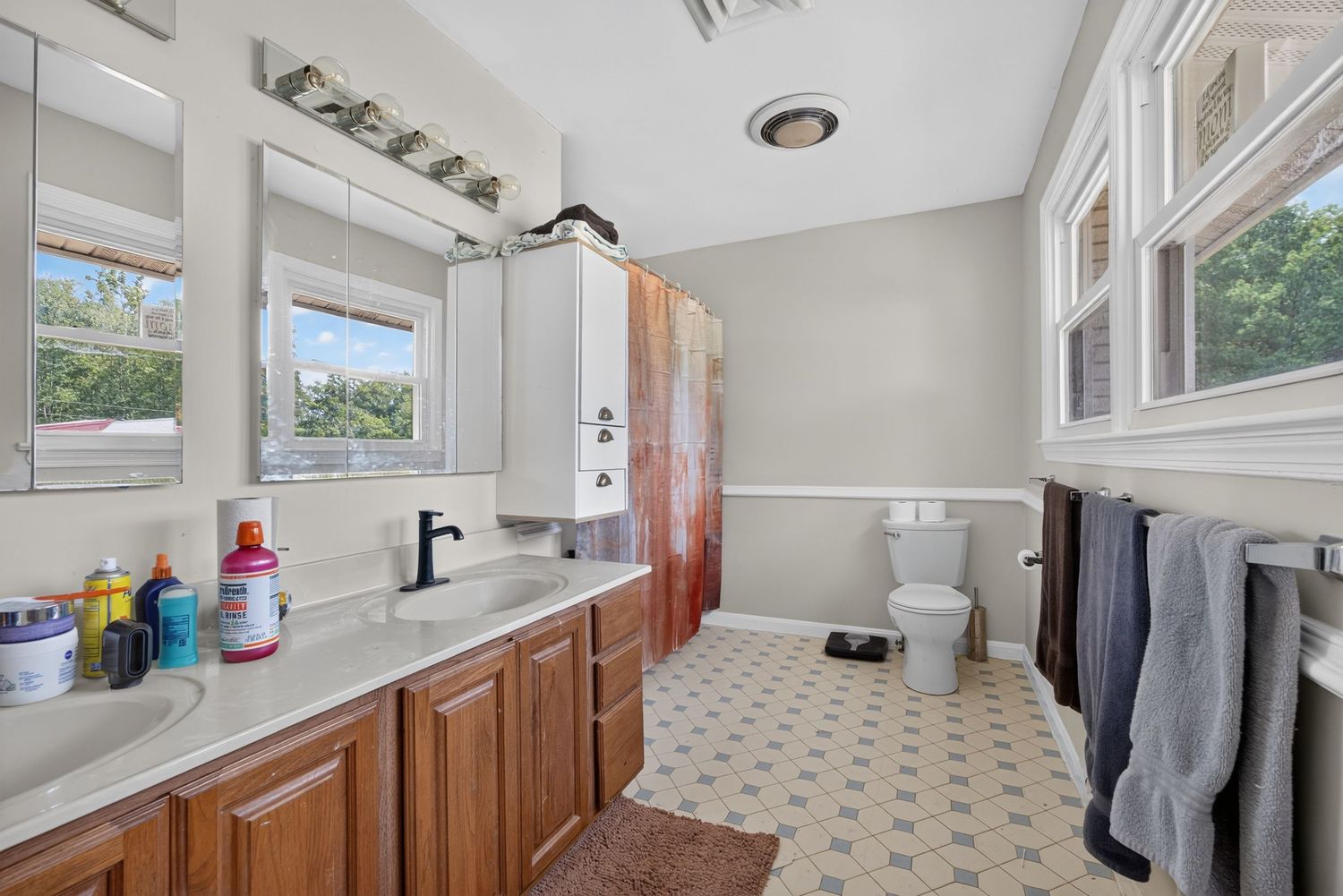Spacious bathroom with double vanity, wood cabinets, patterned tile floor, toilet, and natural light from two windows.