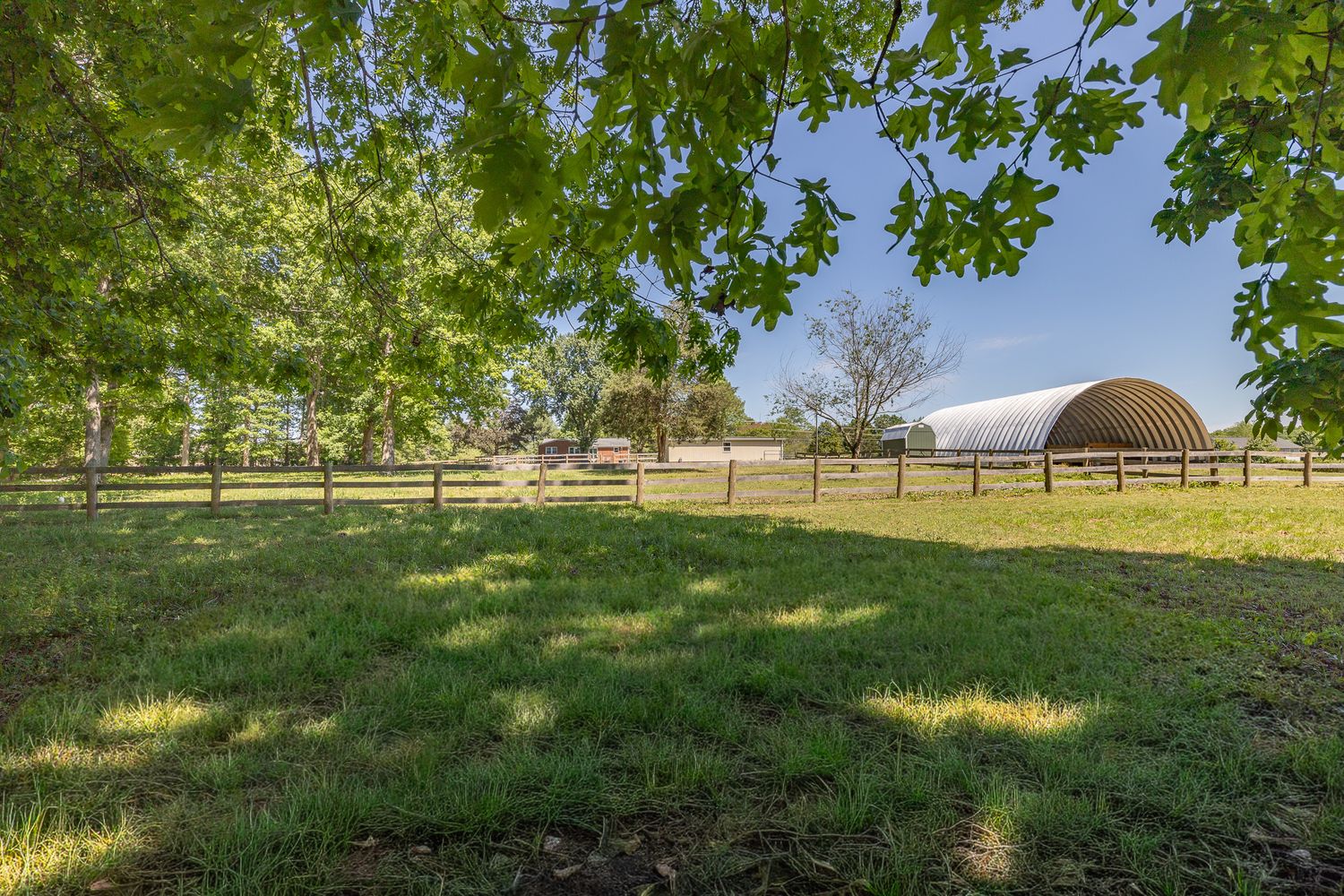 Lush green farm property with mature shade trees, wooden fencing, and a quonset hut barn under a clear blue sky.