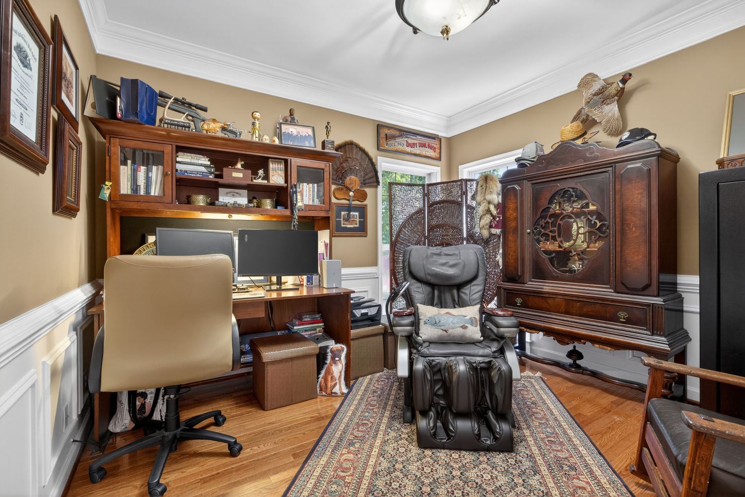 Home office with hardwood floors, built-in shelving, wooden desk, massage chair, and ornate armoire cabinet.