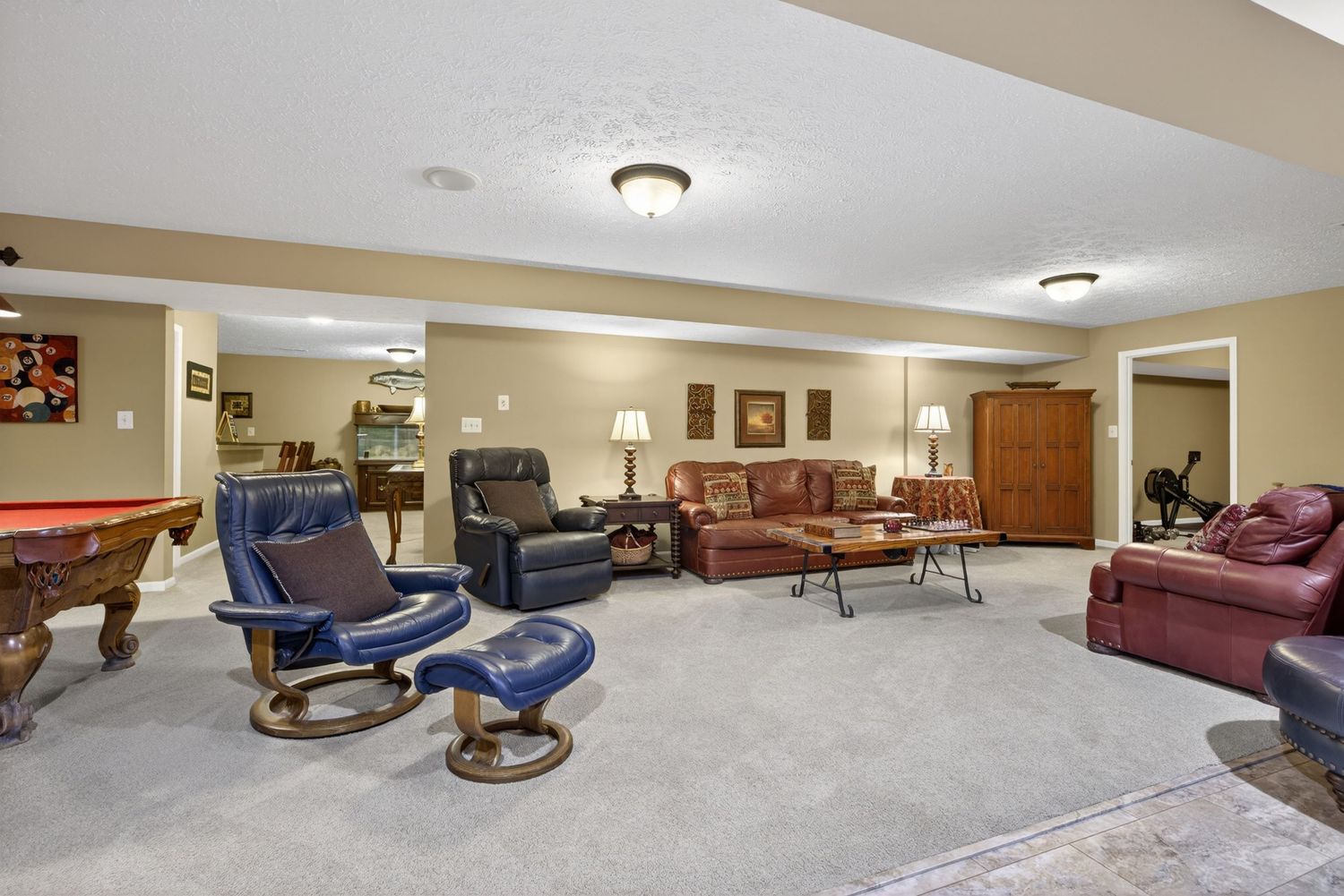 Spacious basement rec room with plush carpet, leather recliners, pool table, and multiple seating areas in warm neutral tones.