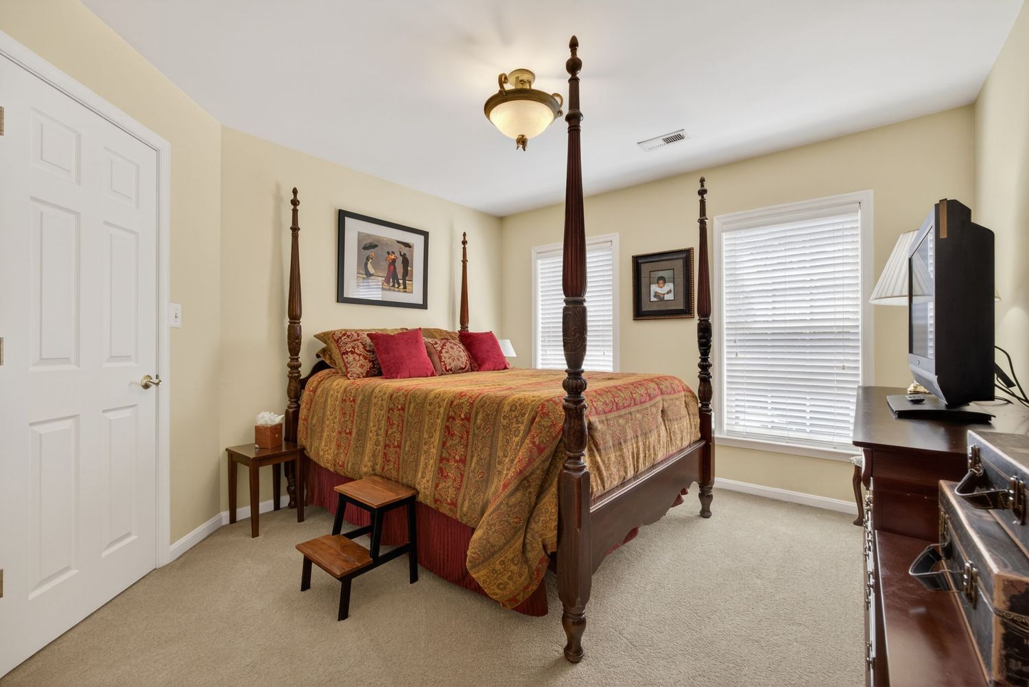Elegant bedroom featuring a dark wood four-poster bed with red bedding, carpet flooring, and a TV on a wooden dresser.