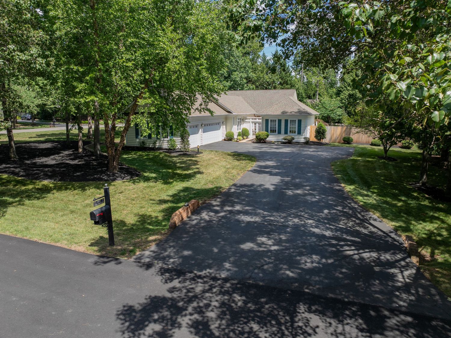 Tree-lined driveway leading to a charming single-story home with white garage door and lush green lawn.