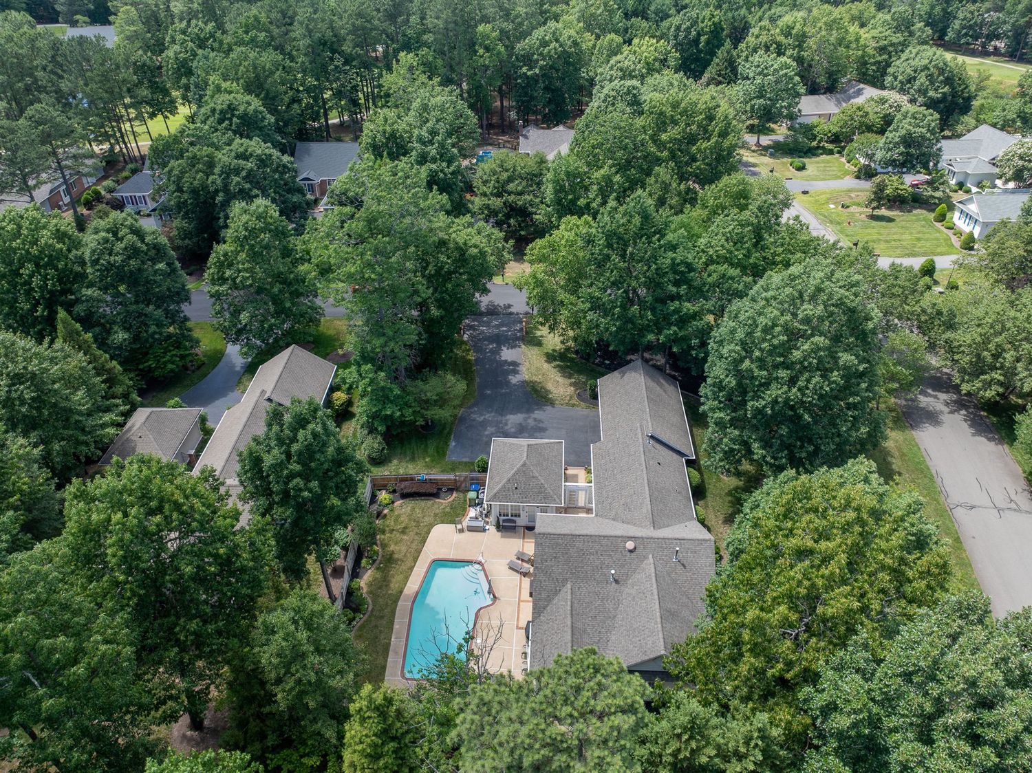 Aerial view of a suburban home with a backyard pool surrounded by lush green trees in a residential neighborhood.