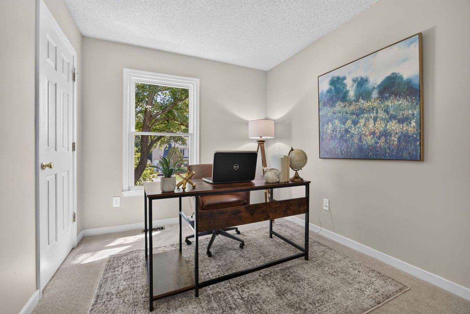 Home office with a dark wood desk, laptop, globe, and a landscape painting above on light gray walls.