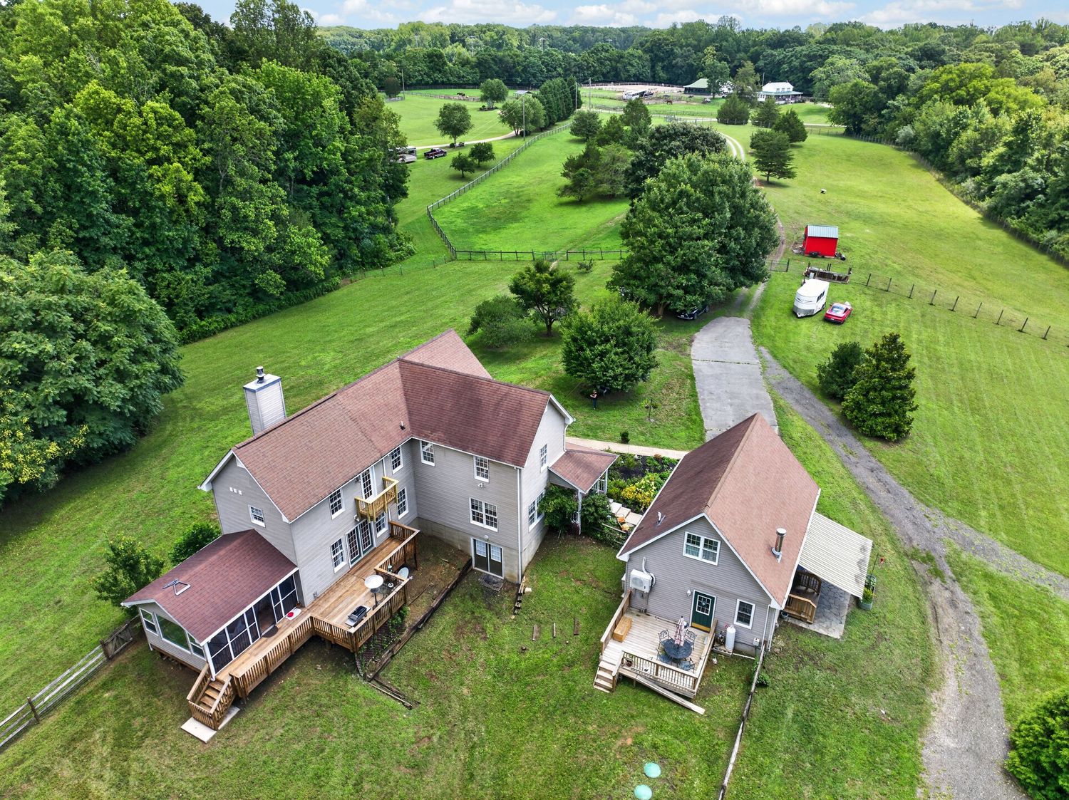 Aerial view of a large farmhouse with deck, surrounded by lush green fields, trees, and a red barn on a rural property.