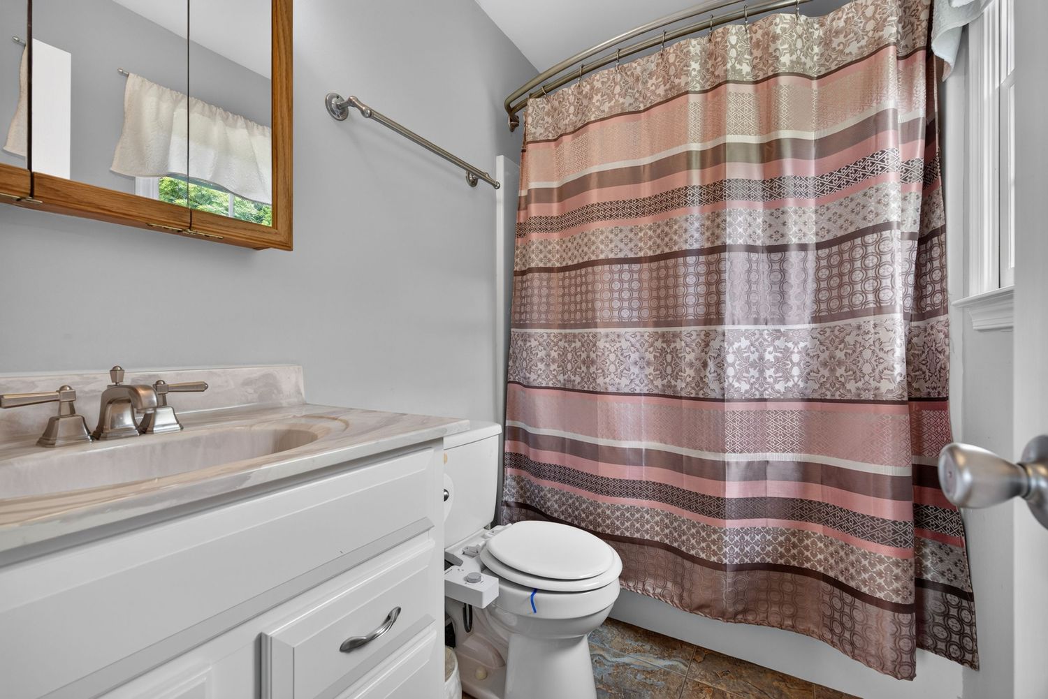 Small bathroom with white vanity, medicine cabinet mirror, toilet, and patterned pink-brown shower curtain on curved rod.
