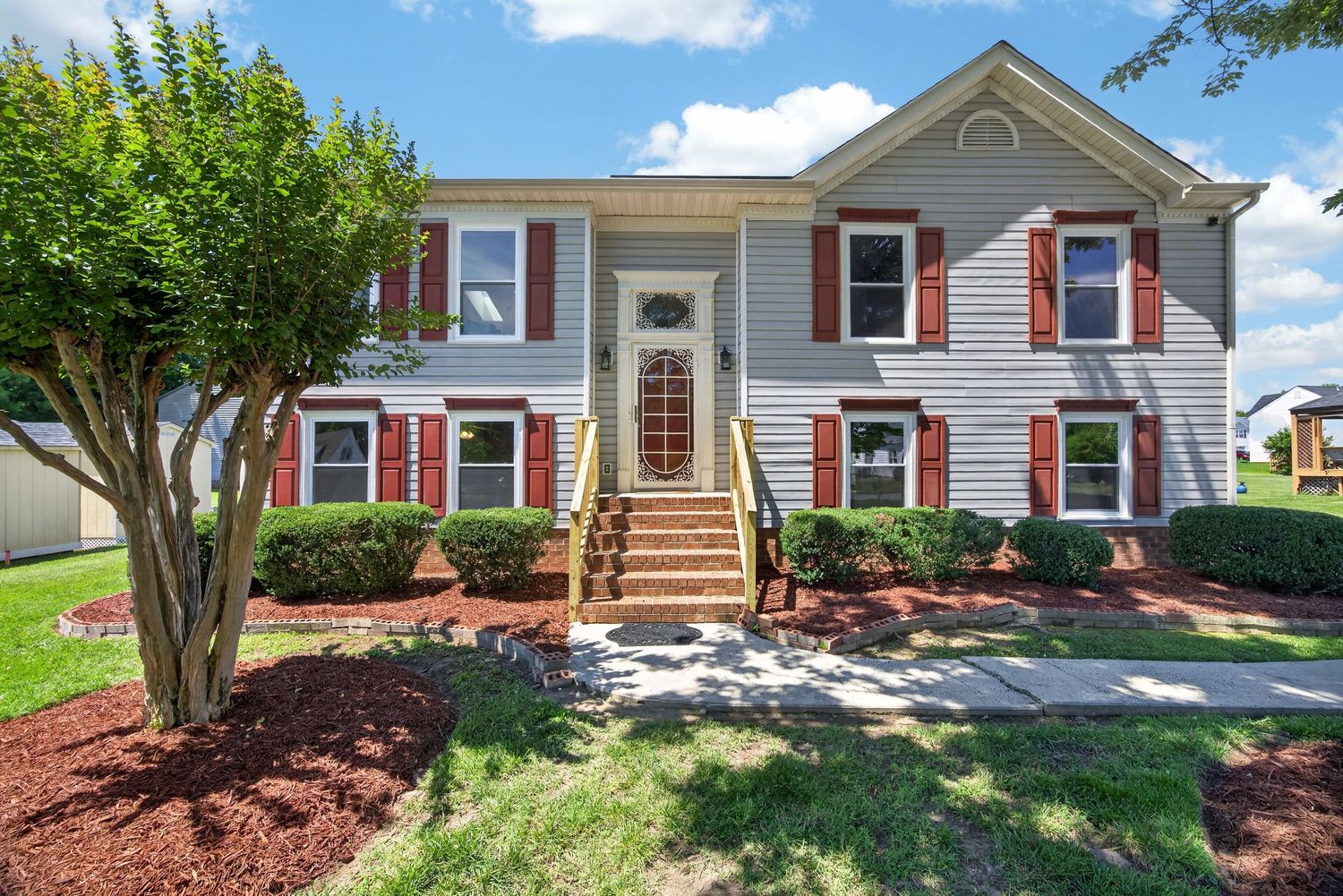 Two-story colonial home with gray siding, red shutters, brick steps, and lush green landscaping on a sunny day.