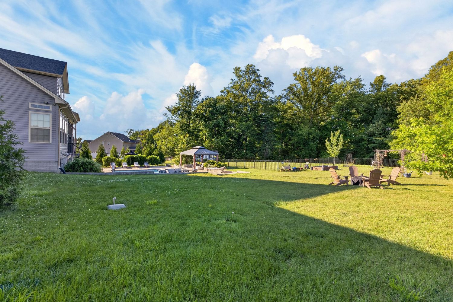 Lush green backyard with lawn chairs, gazebo, and tall trees under a blue sky with white clouds.