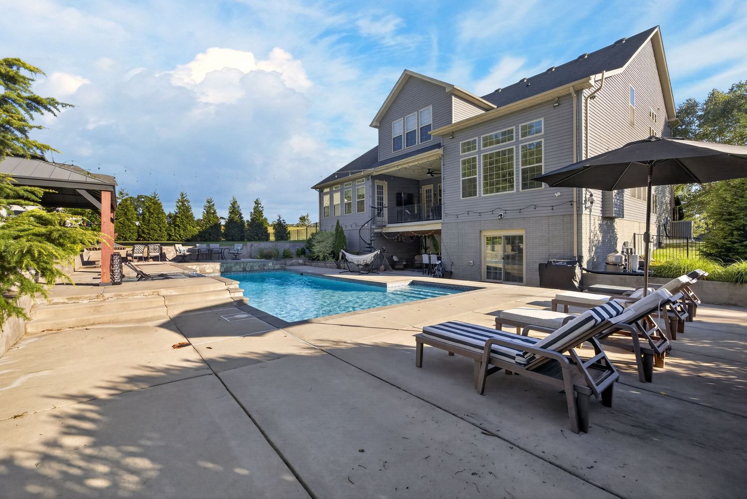 Luxury home with large outdoor pool, lounge chairs, umbrella, and spacious patio surrounded by lush greenery.