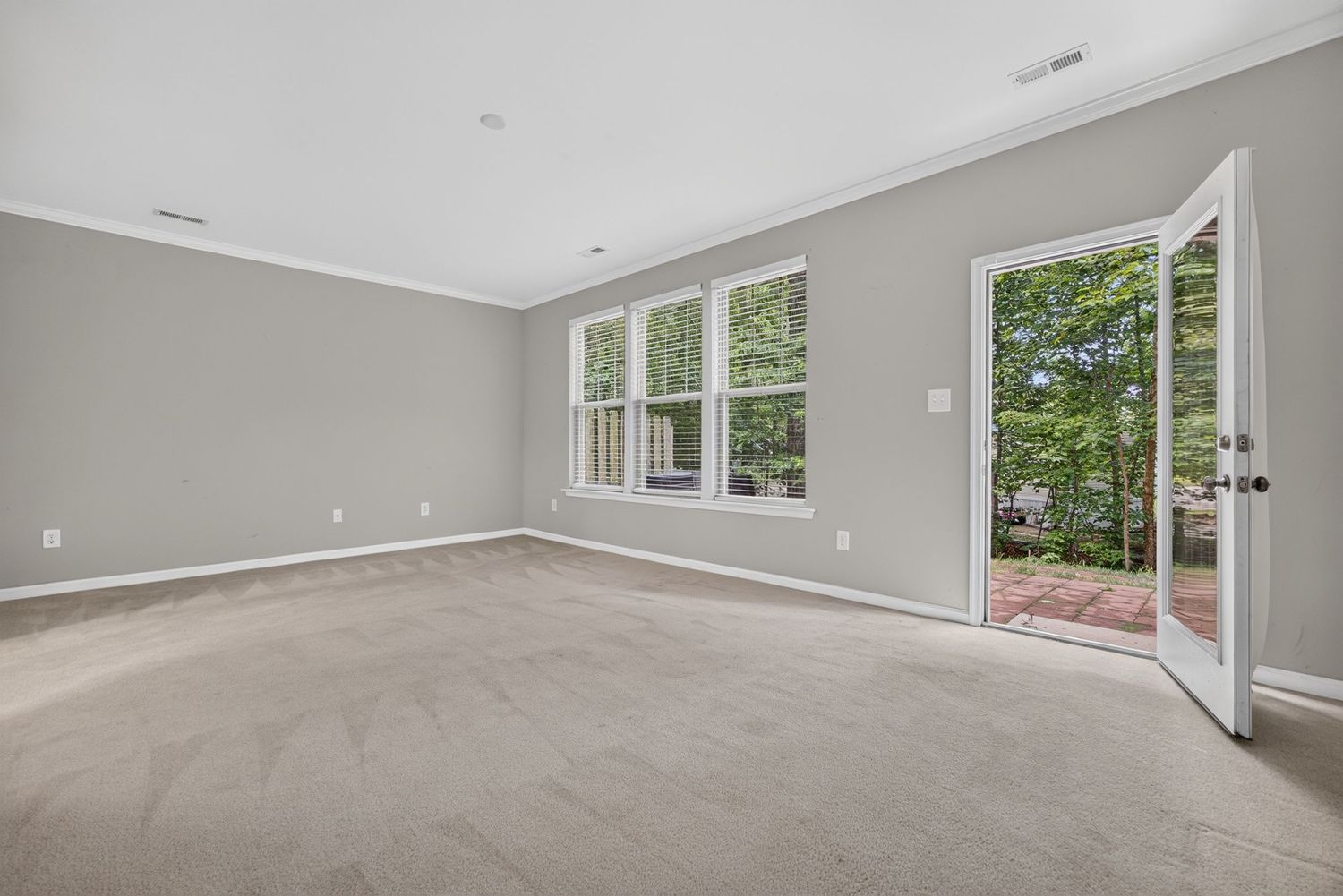 Spacious empty room with light gray walls, plush carpet, crown molding, and French doors opening to outdoor patio.